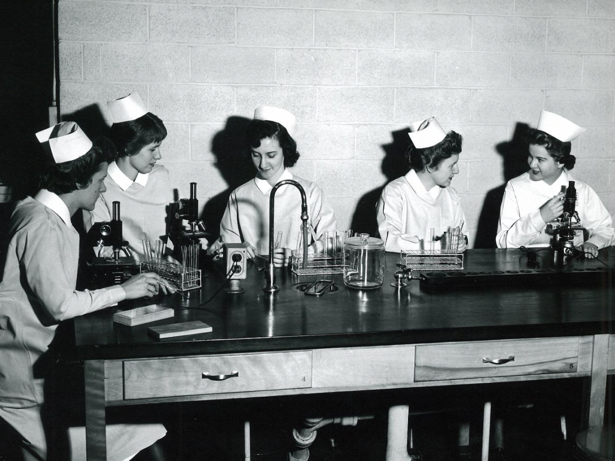 Five nursing students in traditional white uniforms with caps working at a laboratory bench with microscopes and scientific equipment in a black and white photograph.