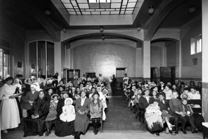 Historic black and white photo of hospital waiting room with nurses attending to families and children in formal attire.