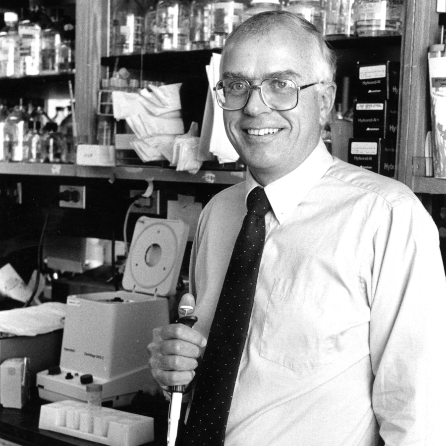 Black and white photograph of a smiling male scientist or researcher in shirt and tie holding a pipette in a laboratory, with equipment and specimen containers visible on shelves behind him.