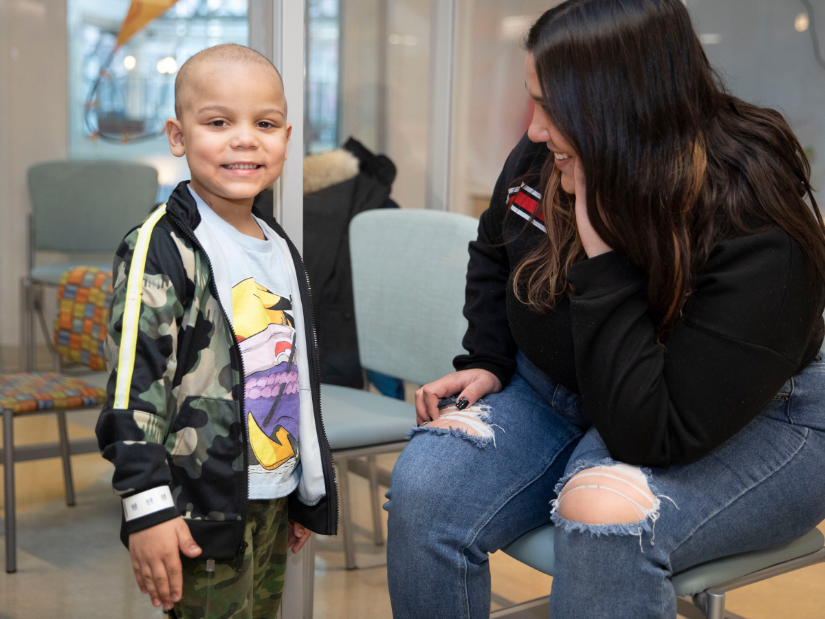 Smiling young child with shaved head in camouflage jacket interacting with a woman in black sweater in what appears to be a hospital waiting area.