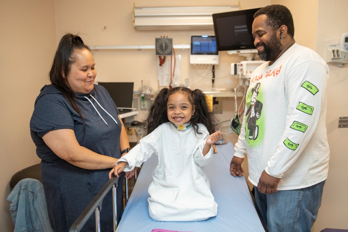 A child sits on a hospital bed while two caregivers stand beside them, holding hands in a medical exam room with monitors and equipment.