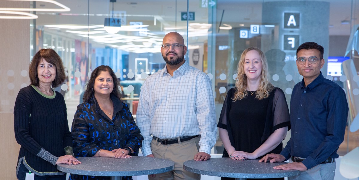 Five adults stand together around tall tables in a bright, modern indoor space, posing as a group in a professional setting.