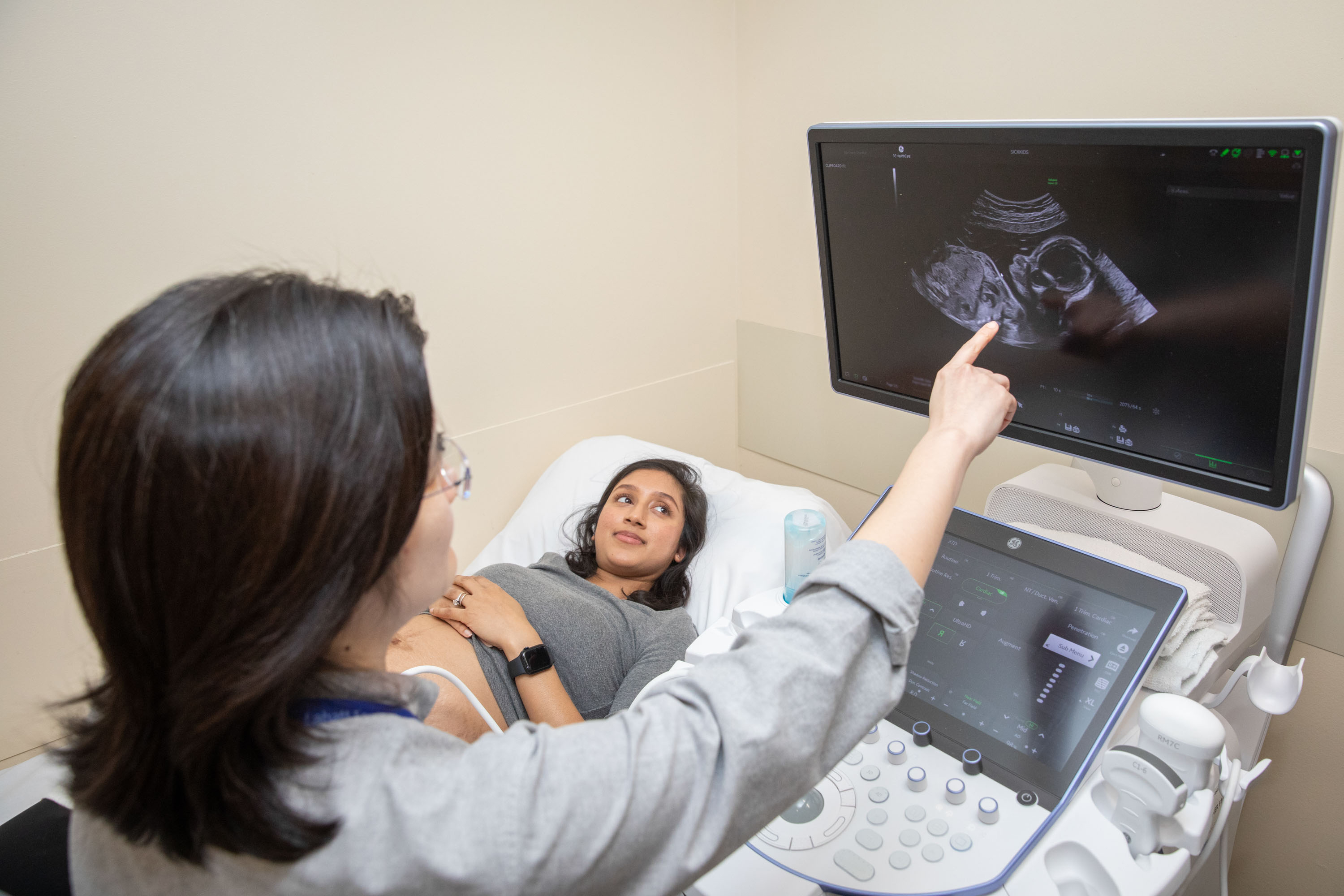 A healthcare professional points to an ultrasound image on a monitor while a patient lies on an exam bed during a scan.