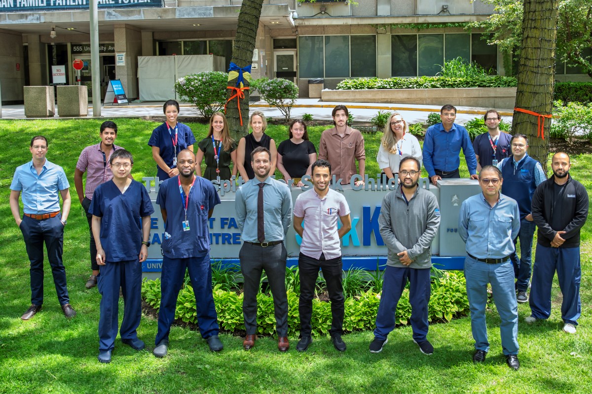 A group of fifteen people standing outdoors on grass in front of a hospital sign, wearing business casual and medical attire.