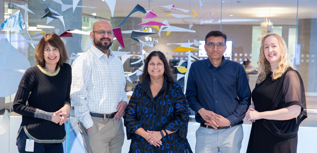 Five healthcare staff stand together in a bright hospital atrium, posing as a team with glass walls and colorful artwork behind them.