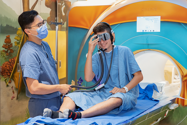 Teen boy looks into virtual reality goggles while seated on an MRI bench. A staff member stands by.