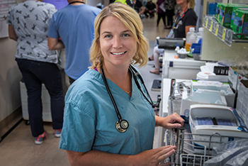 Woman wearing scrubs and a stethoscope stands in a hospital hallway.