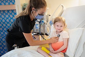 Nurse wearing a medical mask holds a stethoscope to a young girl's chest. The girl sits smiling on a hospital bed.