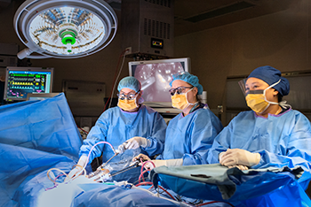 Three people dressed in medical gowns, gloves, caps and goggles stand at a surgical bed in an operating room.