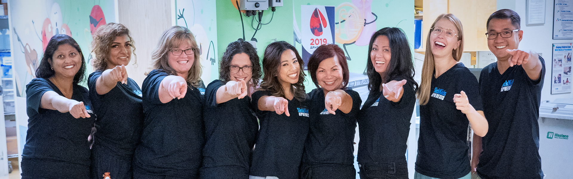 Nine people stand together smiling, pointing at the camera.