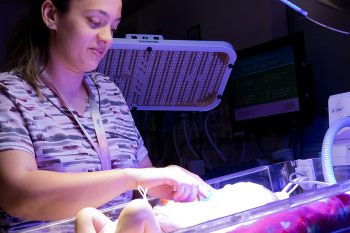 Woman wearing scrubs stands over an infant in a hospital crib.