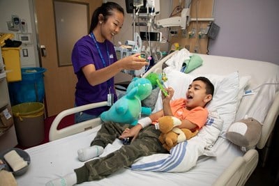 Teenager holds a toy, playing with a child in a hospital bed.