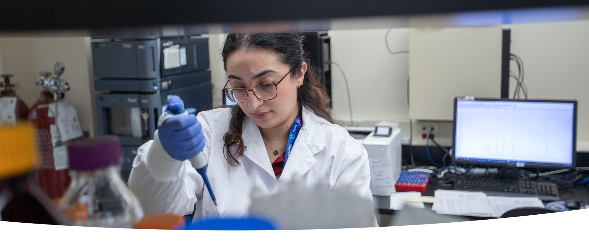 A lab professional wearing a lab coat and blue gloves using a large pipette. In the background is various lab equipment and a computer screen.