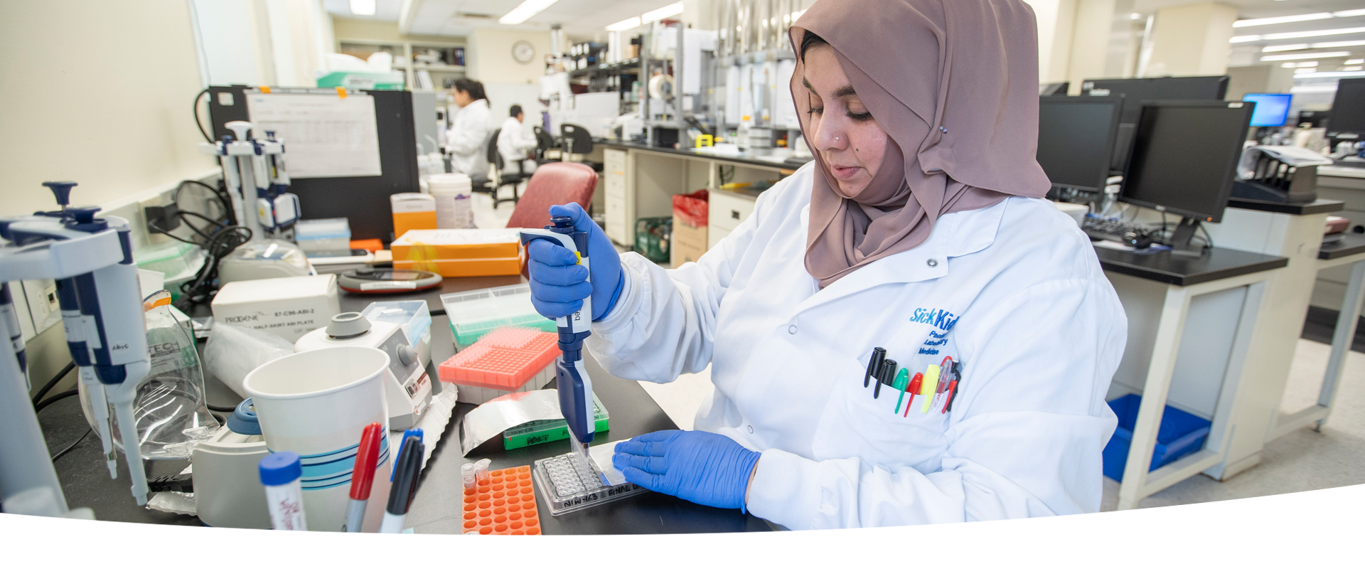 A laboratory professional pipetting into a multiwell plate at a lab bench. In the background, there are other lab professionals at work.