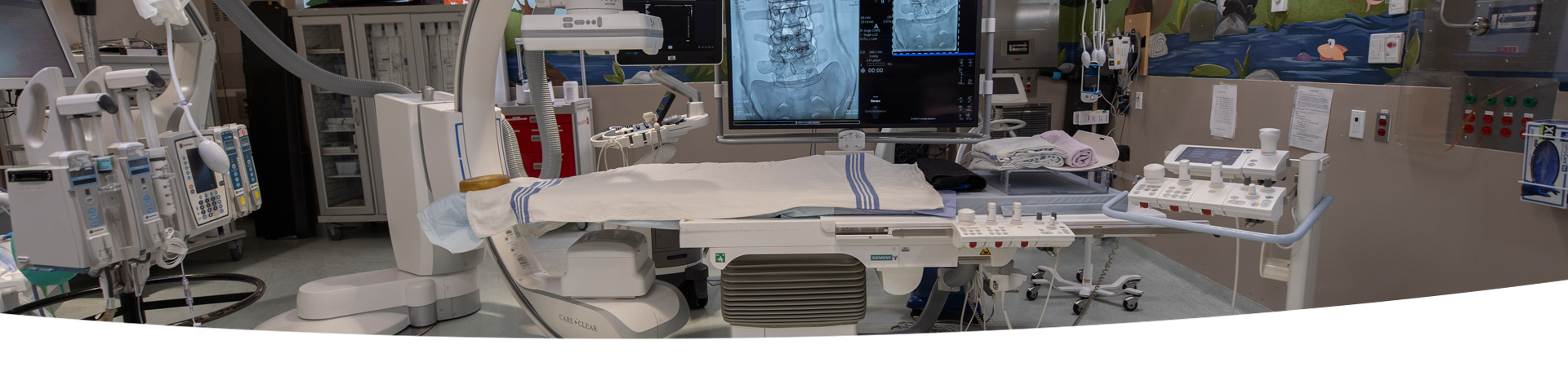 A room with a Single Plane Digital Angiography unit. There is a  variety of hospital equipment in the background.