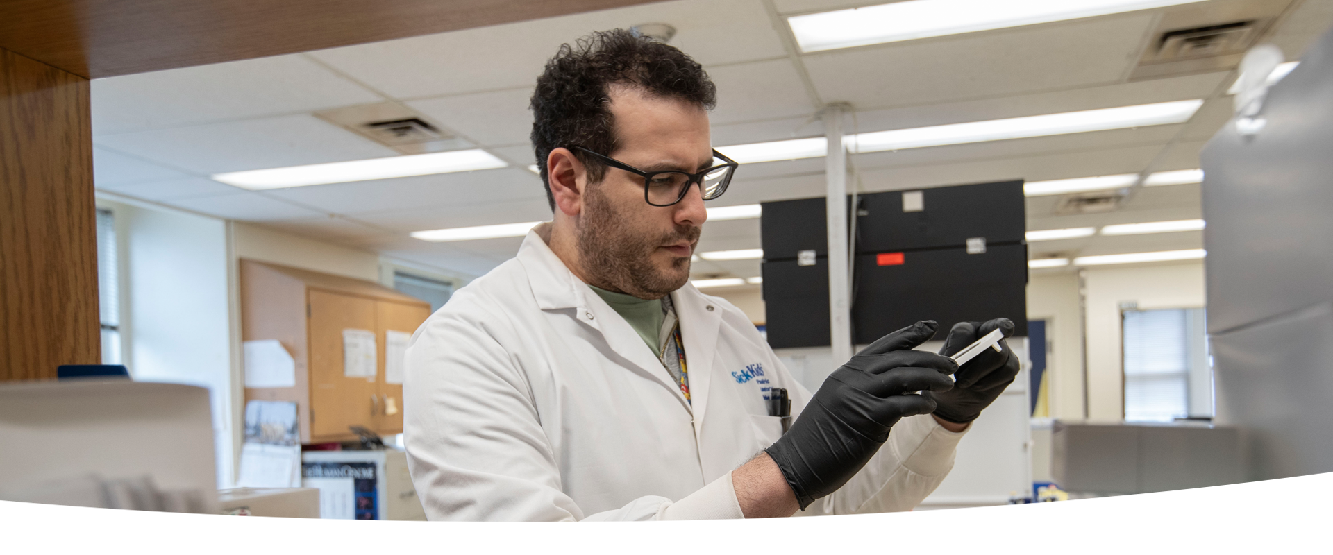A lab professional wearing a lab coat and black gloves is examining a piece of equipment in his hands.