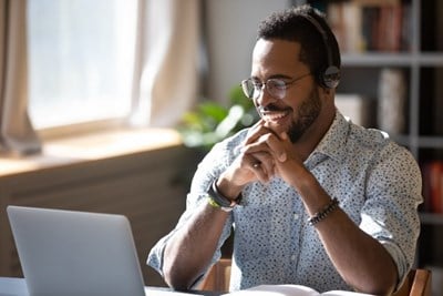 A man wearing a headset sits at a desk with a laptop and book open in front of him