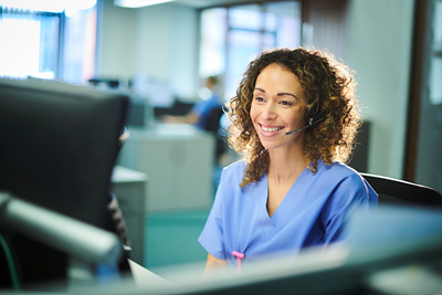 Woman wearing headset and hospital scrubs, seated at a desk.