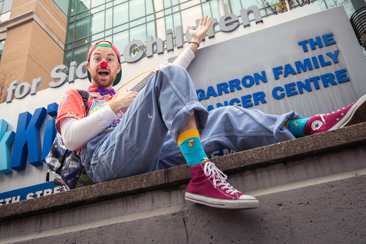 Jazz, a therapeutic clown, sitting by the SickKids sign by the Atrium entrance with ukelele in hand.