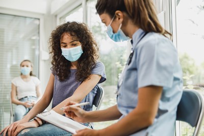 A woman in a mask sits in a chair in conversation with a health-care professional seated in the chair beside her with clipboard and pen in hand.