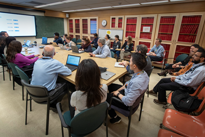 A group of about 20 people sitting around a table in a boardroom. Many of them have their laptops open or looking toward a large screen at the front of the room that is displaying a presentation slide.