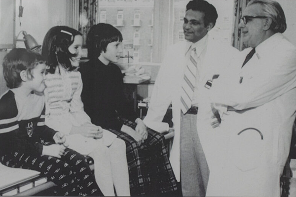 Black and white photo of Dr. Bibudhendra (Amu) Sarkar and Dr. Andrew Sass-Kortsak speaking with three children with Wilson disease