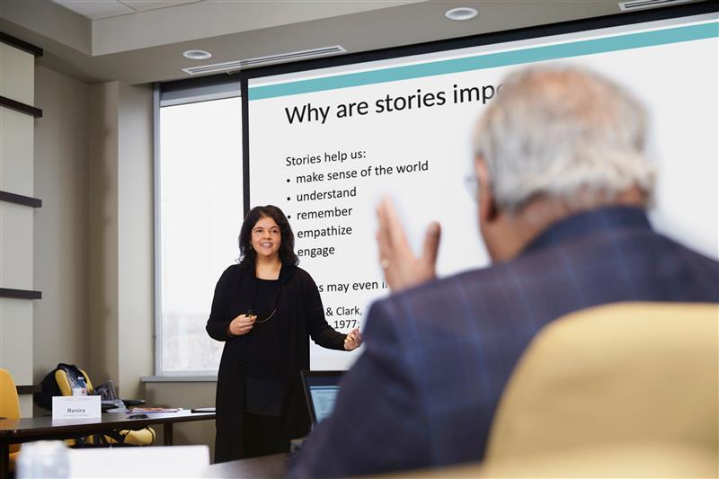 A person presenting in a conference room with a slide titled 'Why are stories important?' listing benefits such as making sense of the world, understanding, remembering, empathizing, and engaging. Another person is seated in the foreground, facing the presenter.