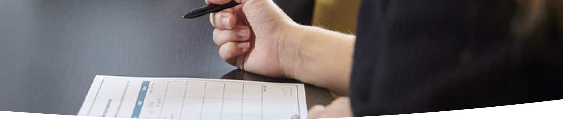 A person wearing a dark long-sleeve shirt is writing with a pen on a grid-layout form or worksheet placed on a dark table. Only the hand and part of the arm are visible.