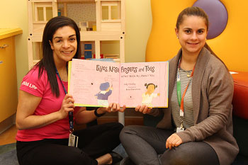 Two women wearing SickKids lanyards hold up a picture book 