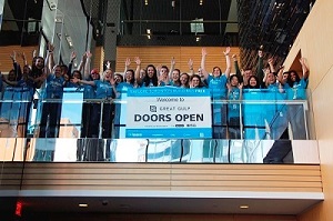 A group of volunteers wearing blue shirts wave and lean over a railing with a banner that reads Welcome to Doors Open