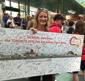 A woman holds a large banner covered in signatures. The banner reads 'SickKids welcomes the Toronto 2015 Pan Am Games Torch Relay" the