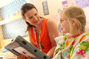A woman wearing a SickKids lanyard sits and talks with a young girl in a hospital room