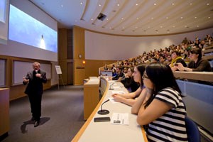 Man stands at the front of a lecture hall filled with people.