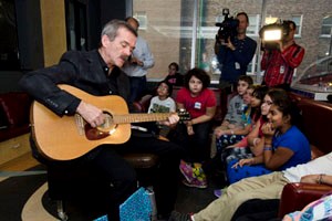 Man plays guitar with children seated around and news cameras in the background.