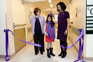 A young girl wearing a purple scarf cuts a large purple ribbon. Two women also dressed in purple stand next to her smiling