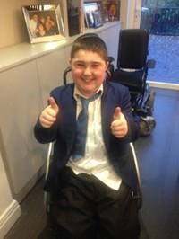 Boy seated in chair wearing jacket and tie holding his thumbs up.