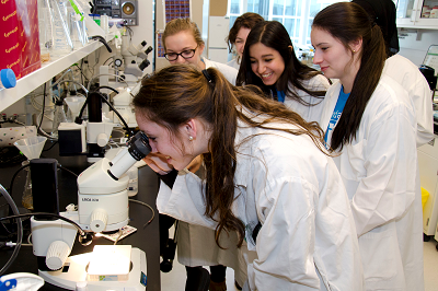 In a lab, a group of teenage girls wearing lab coats observe as another teen looks through a microscope