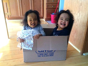 Two young twin children smile while sitting inside a cardboard box