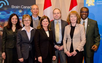 A group of 7 adults stand in front of canadian flags and banners that say Brain Canada and Canadian Cancer Society