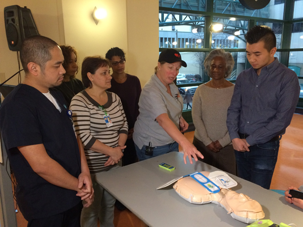 A group of 6 adults watch attentively as an instructor speaks while referencing a CPR dummy