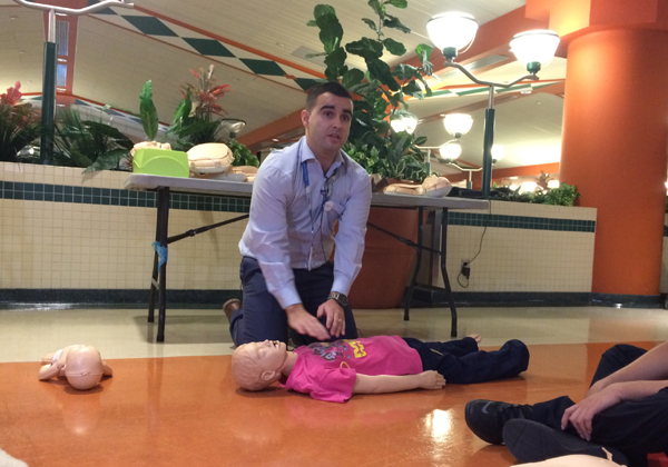 A man wearing a SickKids lanyard kneels and does chest compressions on a CPR Child dummy