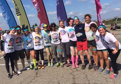 A group of 12 adults wearing athletic clothes covered in colourful pigments huddle together outside