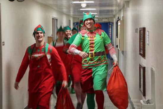 Men dressed as elves smile as they walk down a SickKids hallway carrying bags of toys