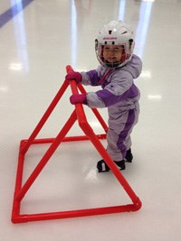 A young girl in a snowsuit, skates and helmet leans against a supportive device on an ice rink