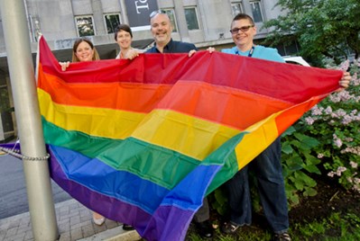 A group of four people standing at the bottom of a flag pole holding the Pride Flag at SickKids