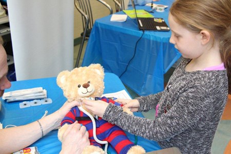 A young girl holds a medical thermometer against a teddy bear