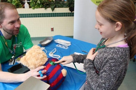 A young girl holds a stethoscope against a teddy bear, while talking to a man wearing a SickKids Child Life tshirt