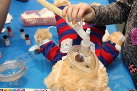 A teddy bear lies on a table while a child's hand holds an anesthesia mask against its mouth