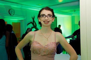 A teen girl dressed in formal wear smiling. Behind her there are colourful lights and silhouettes of people dancing