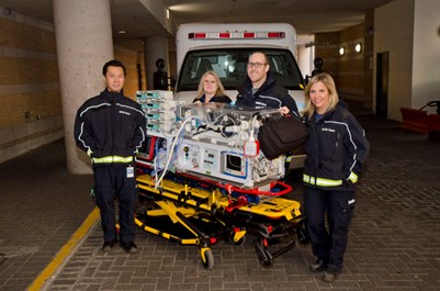Four people in matching coats and pants stand by an incubator with an ambulance behind them.